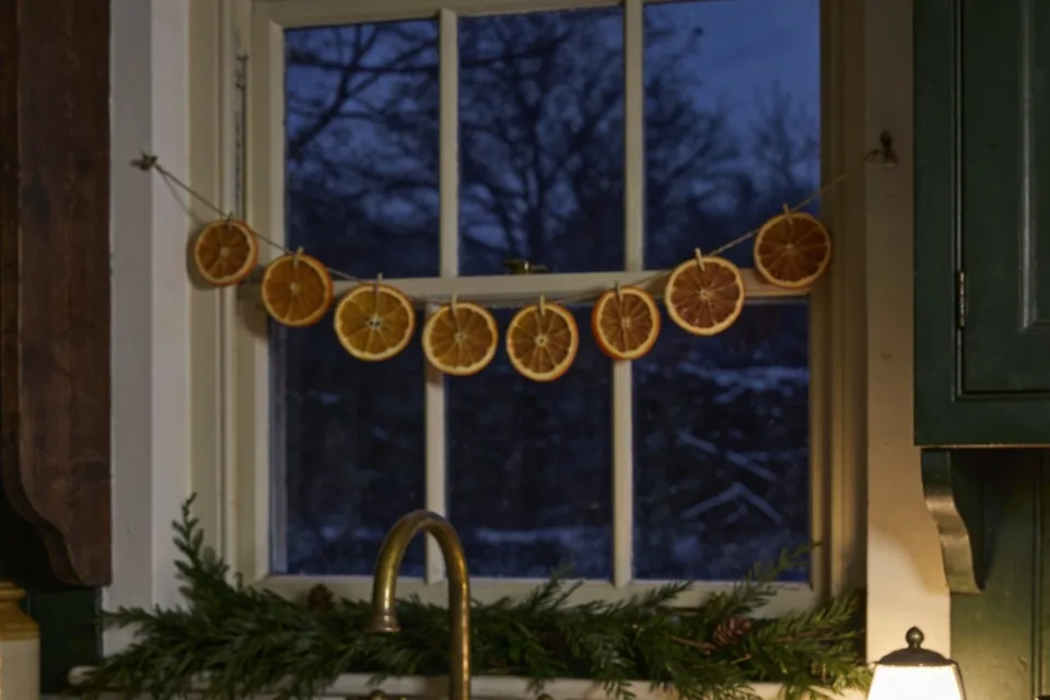 Eight dried orange slices pegged on a twine string across a white painted sash window at winter dusk, bare trees and snow outside, cedar boughs with a few small cones laid along the sill behind an old brass tap, the edge of a dark wood dresser at the left and a green painted cupboard and the shade of a small lit lamp at the right