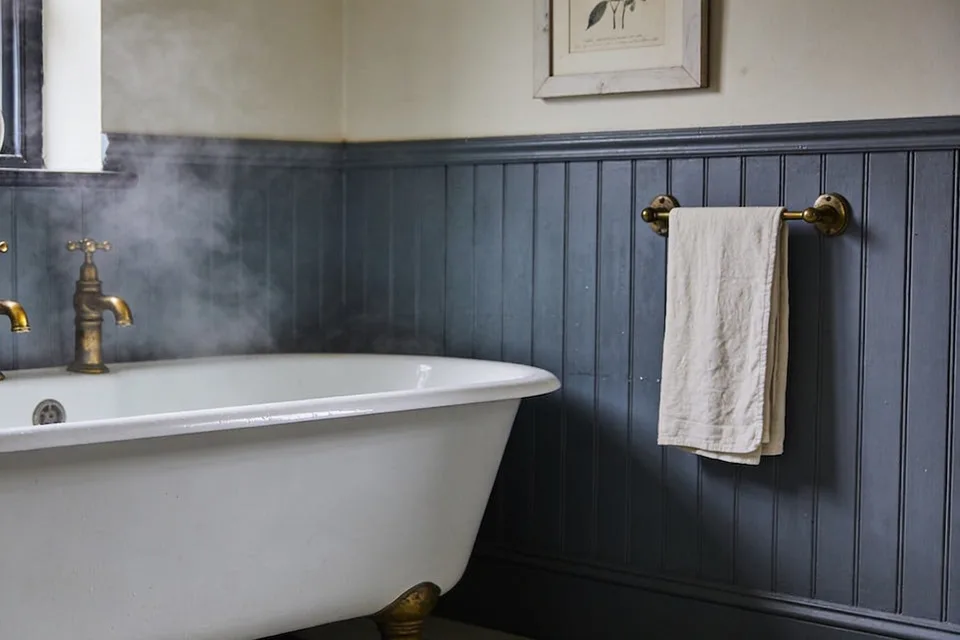 Narrow beaded boards painted a dark slate blue running up a bathroom wall to a moulded rail of the same blue, cream plaster above with the lower edge of a white-framed botanical print, a worn linen hand towel hanging from an old brass towel bar on the boards, and the rim of a white roll-top bath with two brass taps on the left, a little steam rising beside them