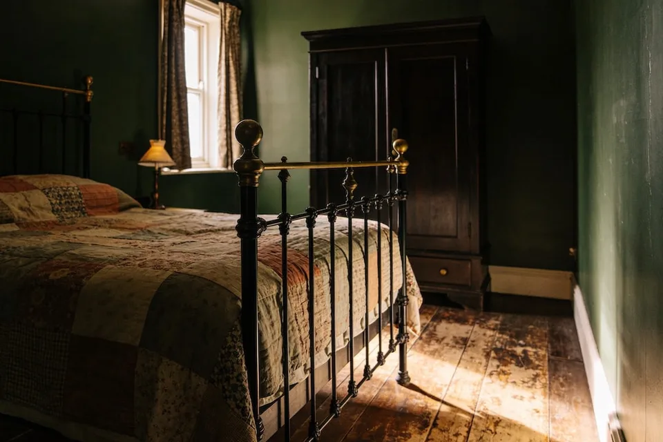 A small cottage bedroom in low morning sun with deep green walls, a black iron bed with brass rails and knobs, a faded patchwork quilt in rose, cream, blue and green over the bed, a small lamp with a pleated shade on the windowsill, patterned curtains at a white sash window, a tall dark wooden wardrobe with a drawer at the bottom, a white skirting board, and wide, worn floorboards lit by a patch of sunlight