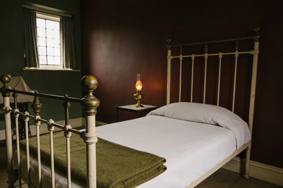 A small guest bedroom with two wall colours: a deep green wall with a small leaded window and checked curtains on the left, and a deep brownish red wall behind a cream-painted iron bed with brass knobs, white sheets and a folded olive green wool blanket at its foot, a small brass oil lamp lit on a narrow table, and an old sewing machine under a cover by the window