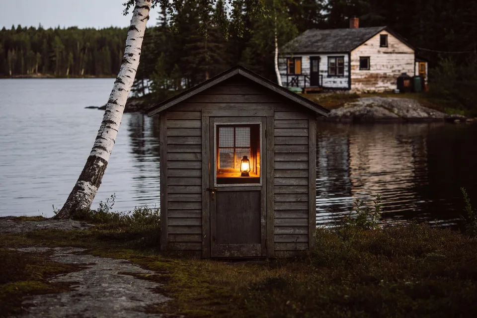 A small one-room bunkie of bare weathered grey clapboard with a peaked roof standing on moss and rock at the water's edge at dusk, seen from the worn path, its plank door shut and an oil lantern burning behind the checked curtain of the window in the door, a leaning white birch crossing the left of the frame, a flat grey lake and a dark spruce treeline beyond, and the weathered white cottage it belongs to on the rocky point behind it to the right
