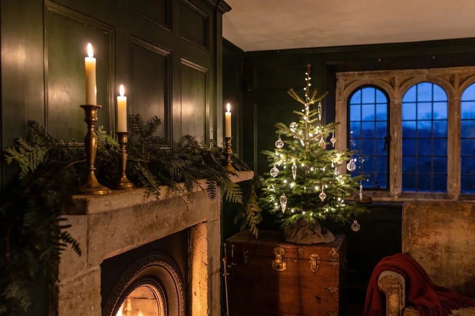 A deep green panelled cottage sitting room at December dusk — fresh fir and fern laid along the stone mantel under three lit brass candlesticks, a fire in the grate below, and a small real tree strung with warm lights and clear glass ornaments standing on an old wooden trunk before an arched leaded window gone deep blue