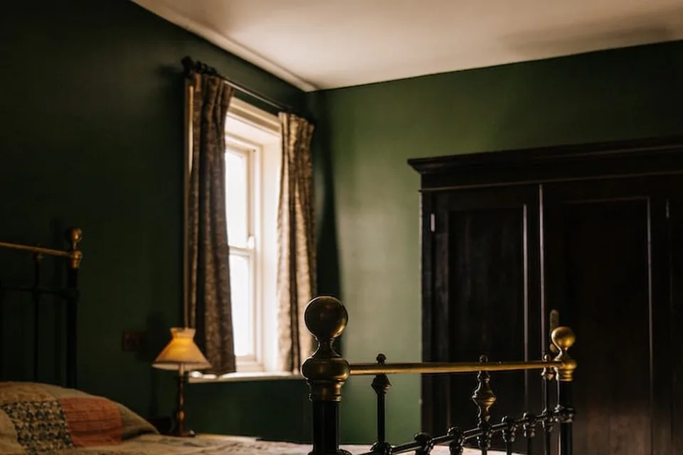 A cottage bedroom in afternoon light with dark green walls, a white sash window hung with a pair of patterned brown and cream curtains on a plain dark iron pole that runs past the frame on both sides, the curtains drawn back to the edges and falling to just below the sill, a small lamp with a pleated cream shade lit beside the window, a dark wooden wardrobe on the right and the brass-knobbed foot of an iron bed in the foreground