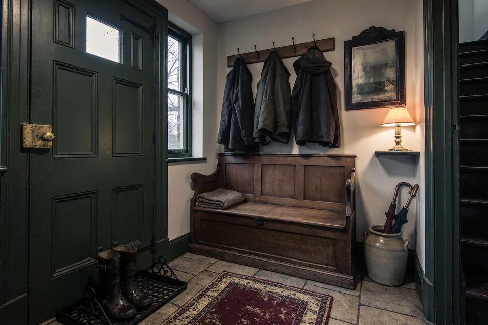 A cottage front entryway on a grey winter afternoon with pale plaster walls and deep moss green painted woodwork, a heavy panelled front door standing open at the left with a small glazed pane and a worn brass knob on a brass plate, a small paned window beside it showing bare trees, a plain wooden peg rail on the far wall hung with three heavy work coats hanging flat, a long oak box settle below it with a panelled back, shaped arms and a hinged lidded seat with a small keyhole, a folded grey wool blanket at one end, a dark framed old mirror on the right wall with heavily foxed and clouded glass, a lit brass table lamp with a cream shade on a narrow shelf beneath it throwing a warm pool of light, a grey salt-glazed stoneware crock in the corner holding two furled umbrellas, a black cast iron boot tray just inside the door with a pair of wet dark rubber boots standing in it, worn stone flagstones underfoot with a faded dark red and cream flat-woven runner laid across them, and a dark wooden staircase rising at the far right