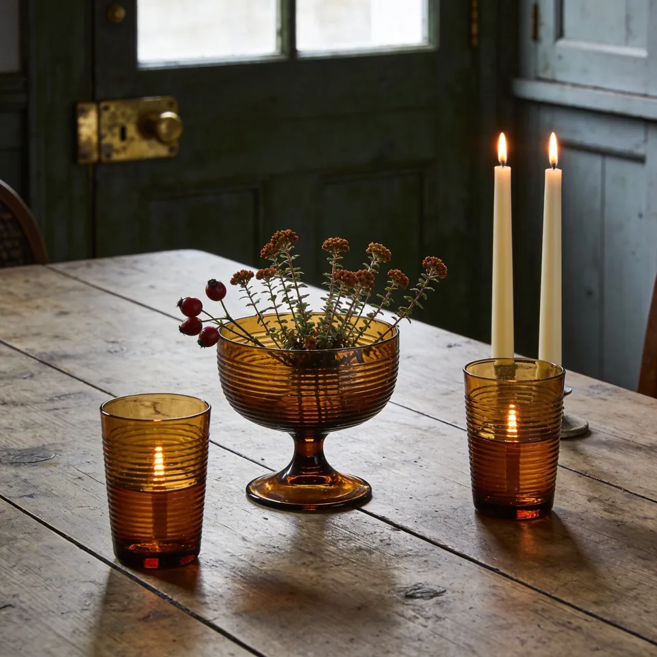 Two ribbed amber glass tumblers glowing with tealights and a footed amber compote holding short stems of rust sedum and rosehips on a bare scrubbed oak table, two lit ivory tapers standing behind and a green-painted door beyond