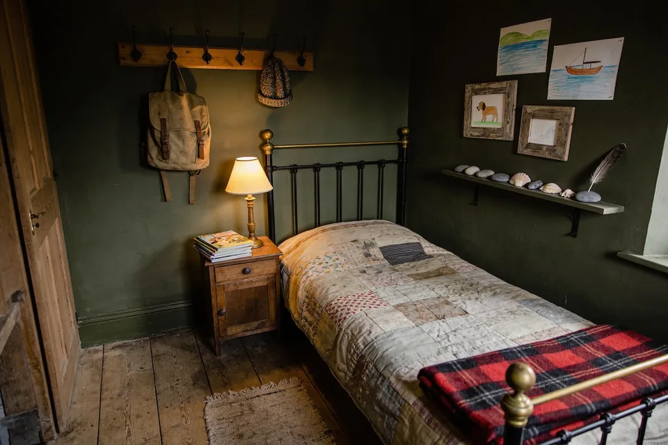 A child's bedroom in an old cottage in the evening, walls painted deep olive green, a black iron single bed with brass knobs made up with a faded patchwork quilt and a folded red and black plaid wool blanket at its foot, a small brass lamp with a cream shade lit on a wooden bedside cabinet beside a stack of picture books, a canvas satchel and a knitted hat hanging from a wooden board of black iron hooks, children's drawings of a lake, a sailing boat and a dog on the far wall, two of them in weathered wooden frames, a narrow shelf holding smooth grey stones, shells and a feather, and wide worn floorboards with a small rag rug