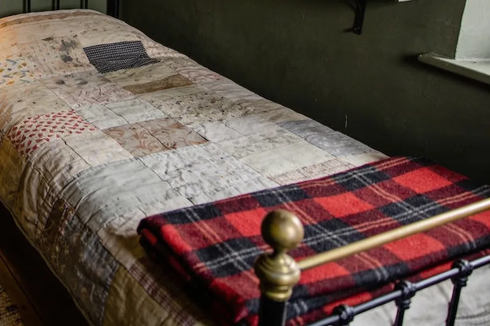 The foot of a narrow single bed made up with a faded patchwork quilt, a folded red and black plaid wool blanket lying across its black iron foot rail with a brass knob and brass top rail, an olive green wall behind and the edge of a white painted window sill at the top right