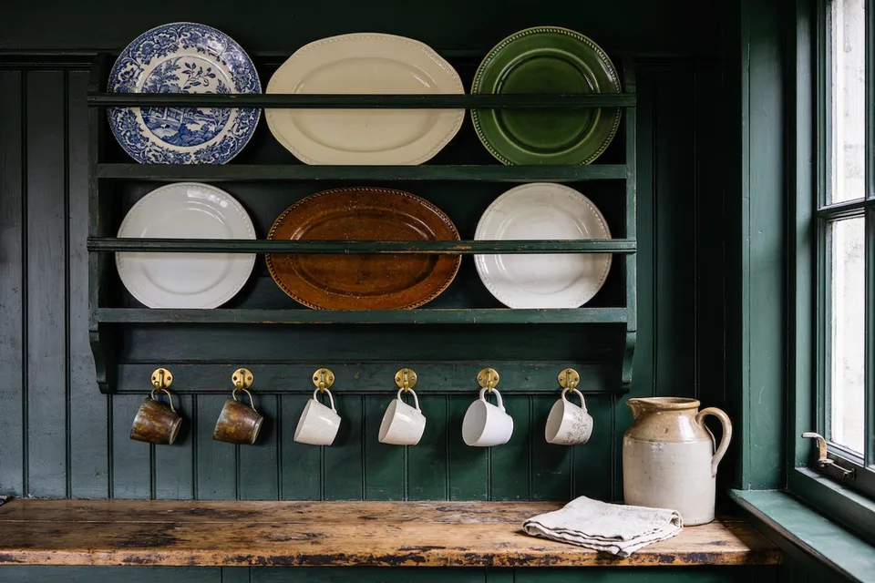 An old dark green painted plate rack on a deep green tongue-and-groove kitchen wall, holding six mismatched plates on edge in two rows: a blue and white transferware plate, a cream oval platter and a green glazed plate above, two plain white plates either side of a brown glazed oval platter below, with six cups hanging from brass hooks along its base, four cream and two brown, over a worn wooden counter holding a cream and brown stoneware jug and a folded linen tea towel, beside a green-framed window