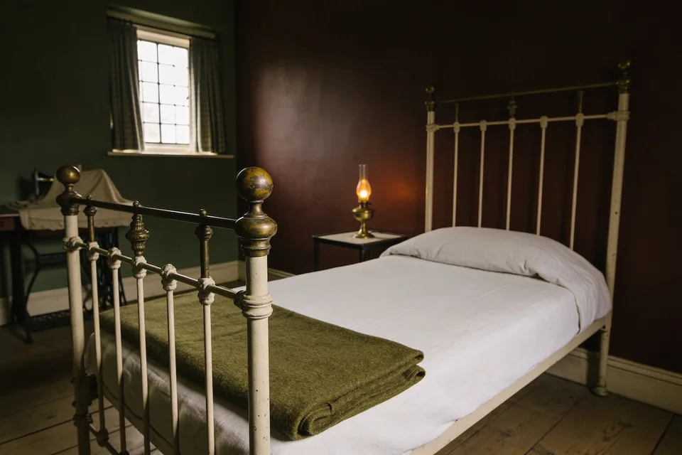 A cream-painted iron bed with brass finials, made up with plain white linen and a folded olive wool blanket laid across its foot, standing against an ox-blood wall on bare floorboards, with a lit brass oil lamp on a small dark bedside table and a treadle sewing machine covered by a cloth in the green-walled corner under a small leaded window