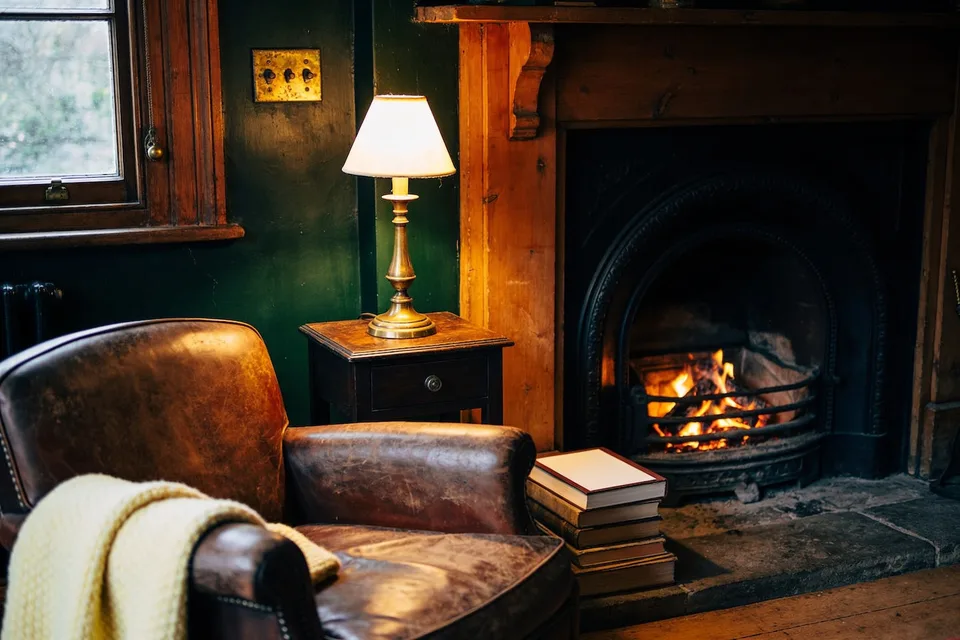 A lived-in English cottage sitting room at night with deep green-black walls, embers glowing in a stone hearth, one lit brass lamp beside a worn leather armchair, a cream wool throw, dark oak and clothbound books in the low warm light