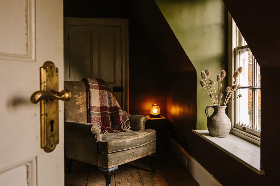 A cottage corner in low amber autumn light — a worn armchair with a plaid wool throw, a small candle lamp lit on the side table, a stone jug of dried teasels on the deep windowsill