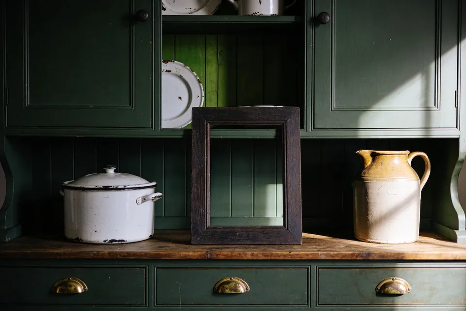 An old dark oak frame standing empty on a green pantry dresser shelf between a chipped white enamel pot and a stoneware jug, the green boards of the dresser showing through it, low warm afternoon light across the shelf