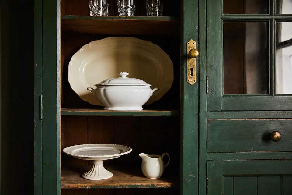 An open bay in a dark green painted kitchen dresser, holding a large scallop-edged cream ironstone platter standing on edge behind a white lidded soup tureen, a footed cream cake stand and a small cream jug on the shelf below, and cut-glass tumblers on the shelf above, with a glazed dresser door and an aged brass knob and keyhole plate at the right