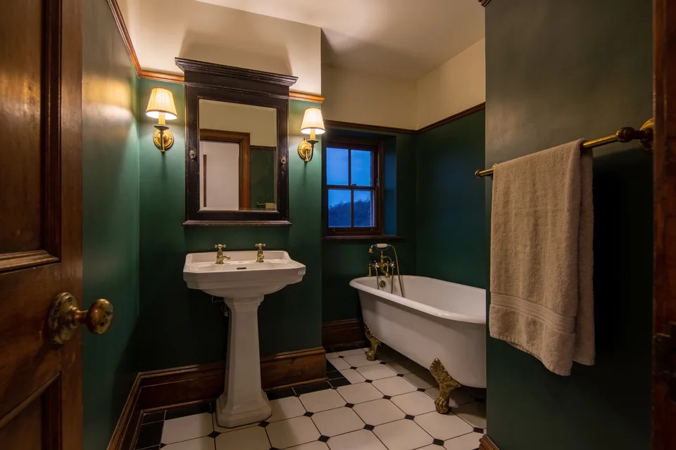 A small bathroom at dusk with deep green walls below a cream upper wall and ceiling, a dark wooden framed mirror over a white pedestal basin with two brass cross-head taps, a small brass wall light with a pleated cream shade lit on each side of the mirror, a white cast iron tub on gilded claw feet with a brass mixer beside a small window showing blue dusk, a beige towel hanging from a brass rail, and white octagonal floor tiles set with small black squares, seen past a panelled wooden door with a worn brass knob