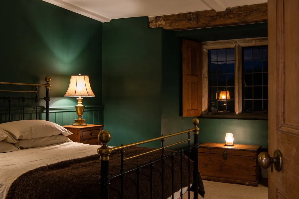 A deep green cottage bedroom at night — a black iron bed with brass knobs dressed in cream linen under a dark brown wool blanket, a lit brass lamp on the pine nightstand, and across the room a small lamp glowing on a wooden chest beneath a leaded stone window gone blue with dusk