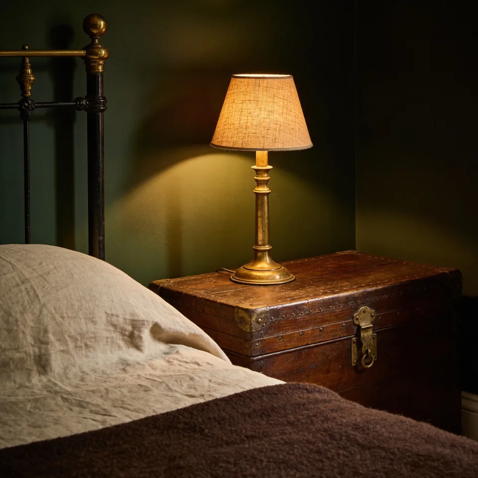 A small brass lamp with a woven linen shade lit on an old brass-latched wooden trunk beside a bed of rumpled natural linen and a dark brown wool blanket, its warm light falling on a deep green wall as the corners go to shadow