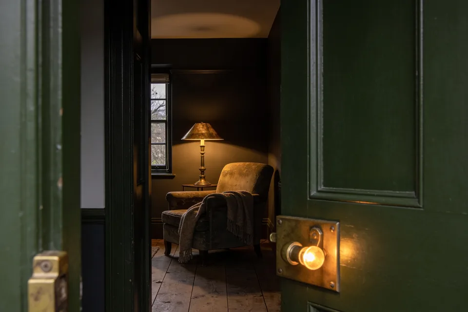 Seen through a deep green doorway, a lamplit sitting room at dusk — a worn armchair with a wool throw beside a lit brass lamp, the door's brass latch catching the light