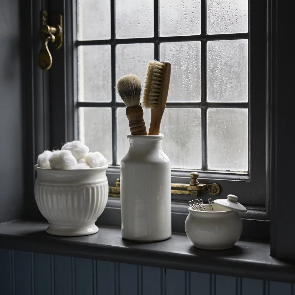 Three white milk glass vessels on a deep painted windowsill — a ribbed footed pot of cotton wool, a taller bottle-shaped jar holding two wooden-handled brushes, a small pot with its lid resting askew over pins — wet gray morning light through fogged small panes with brass window fittings, slate-blue beadboard below