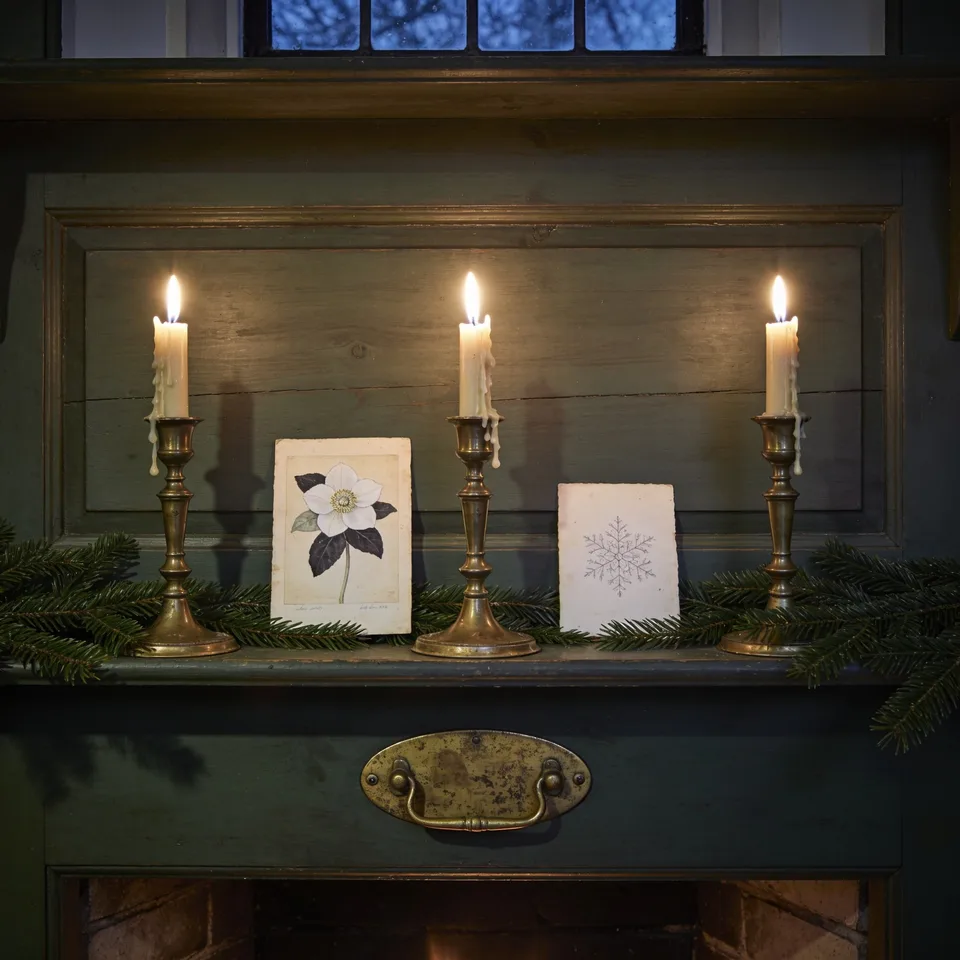 Two prints from the vintage Christmas pack — the Christmas rose and the snow crystal study — propped unframed among fresh fir between three lit brass candlesticks on a cottage mantel at night, wax run down the brass, deep shadow beyond the candlelight