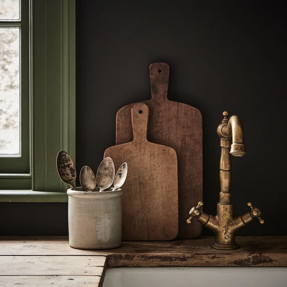 Two handed-down wooden cutting boards leaning against a dark wall beside a stoneware crock of worn spoons and an aged brass tap