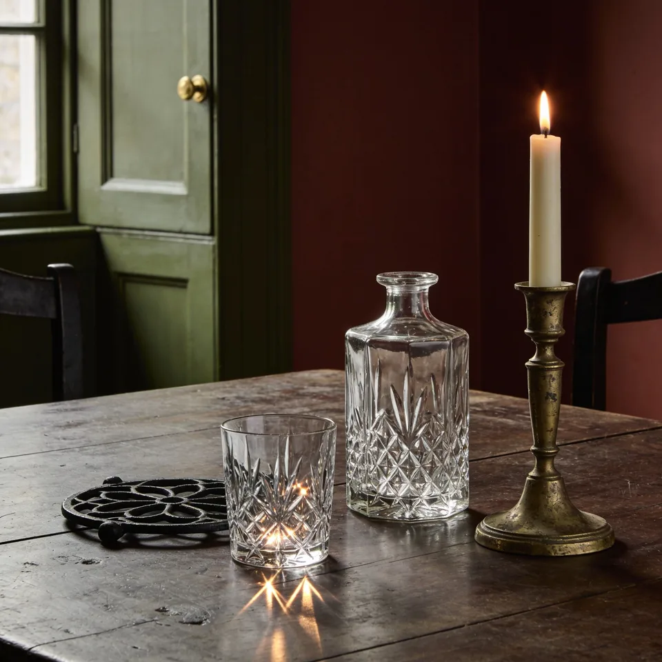 A cut crystal tumbler and an open cut-glass decanter standing on bare worn dark oak beside a black iron trivet, one lit ivory taper in an aged brass candlestick throwing small points of light through the glass onto the wood, a green painted cupboard and deep red-brown wall behind