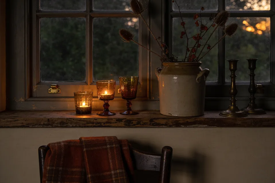 A deep cottage windowsill at dusk, three mismatched amber glass tumblers on the worn oak sill, two holding lit votives, beside a salt-glazed stoneware crock of dried teasels and turning branches, a rust plaid wool blanket folded over the chair below and two darkened brass candlesticks catching the last of the light