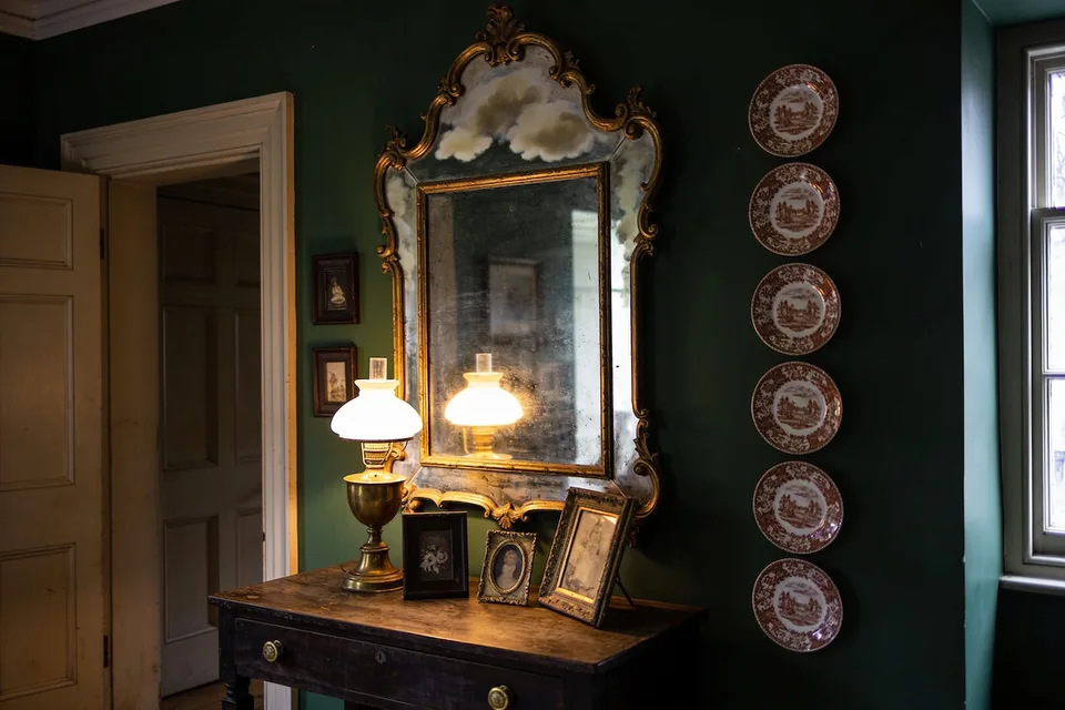 A deep green sitting room wall in low lamplight, a line of brown transferware plates hung beside a bright window, a tall gilt-framed mirror with clouded silvering giving back the glow of a lit brass oil lamp, three small pictures in worn dark and gilt frames standing on the dark wood table below, a doorway open beyond